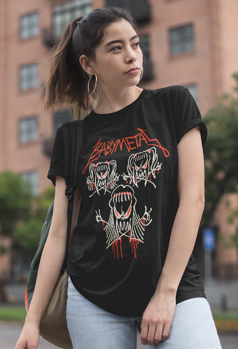 Pode incluir: Uma camiseta preta com um gr&aacute;fico branco de tr&ecirc;s cr&acirc;nios de desenhos animados com o texto "BABYMETAL" em vermelho.