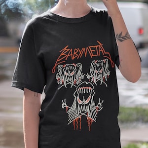 Pode incluir: Camiseta preta com um gr&aacute;fico branco da banda Babymetal. O gr&aacute;fico apresenta tr&ecirc;s cr&acirc;nios de desenhos animados com cabelo comprido e sangue pingando.