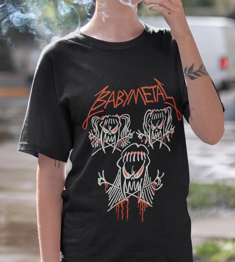Pode incluir: Camiseta preta com um gr&aacute;fico branco da banda Babymetal. O gr&aacute;fico apresenta tr&ecirc;s cr&acirc;nios de desenhos animados com cabelo comprido e sangue pingando.