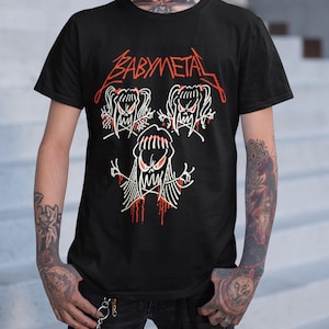 Pode incluir: Camiseta preta com um gr&aacute;fico branco de tr&ecirc;s figuras de desenhos animados com o texto "BABYMETAL" em vermelho acima.