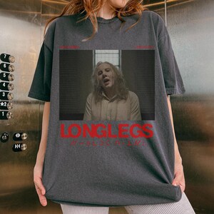 Puede incluir: Una camiseta gris oscura con un gr&aacute;fico de una persona con el pelo largo y el texto "LONGLEGS" en rojo.