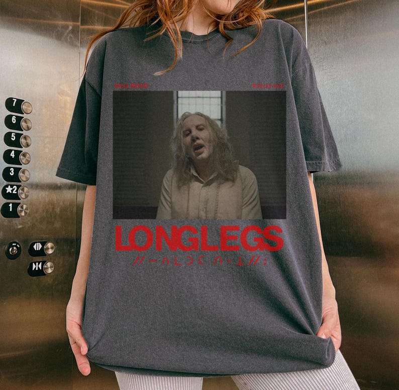 Puede incluir: Una camiseta gris oscura con un gr&aacute;fico de una persona con el pelo largo y el texto "LONGLEGS" en rojo.