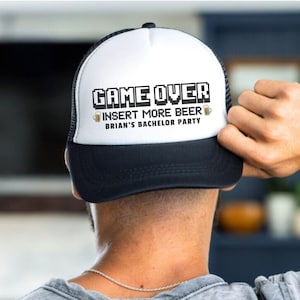 May include: A white and black trucker hat with a pixelated design that says "Game Over Insert More Beer" and "Brian's Bachelor Party".