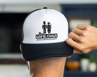 May include: A white and black trucker hat with a black and white illustration of a bride and groom with the text "GAME OVER DAVID'S BACHELOR PARTY" on the front.