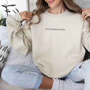May include: A beige sweatshirt with the text "my tummy hurts" printed on the front. The sweatshirt is being worn by a person sitting on a white couch.
