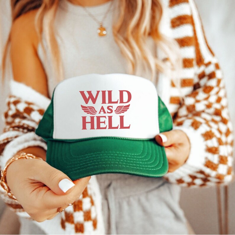 May include: White and green trucker hat with the text "WILD AS HELL" in red lettering with wings on either side of the word "AS".