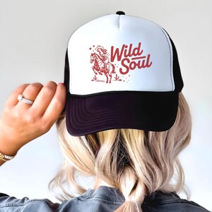 May include: A trucker hat with a white front panel and a black mesh back. The hat features a red graphic of a cowgirl on a horse with the words "Wild Soul" in red script. The brim is black.