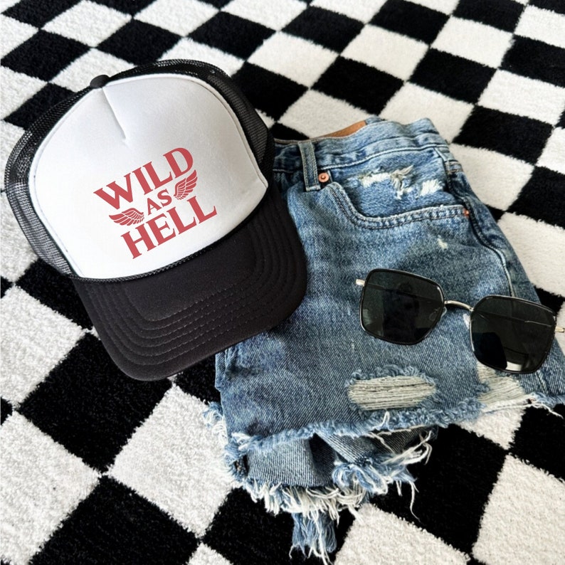 May include: A white trucker hat with a black mesh back. The hat has red text that says "WILD AS HELL" with a red winged design. The hat is on a black and white checkered surface.