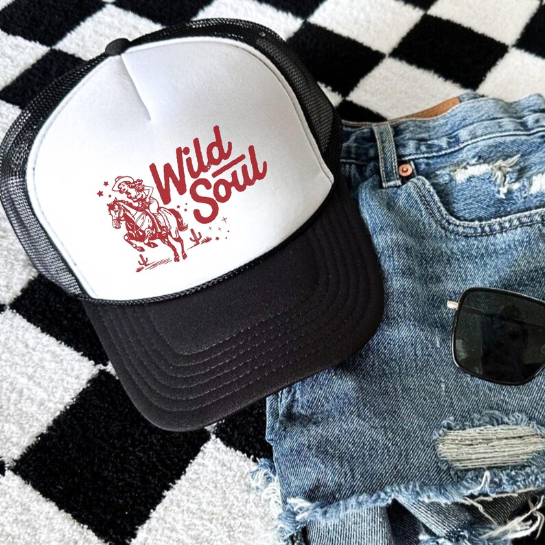 May include: A black and white trucker hat with the words "Wild Soul" in red script, along with a red illustration of a cowgirl riding a horse. The hat is paired with denim shorts and sunglasses.