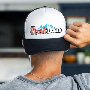 May include: A white and black trucker hat with a blue and white mountain graphic and the text "The Cool Dad" in red and blue lettering.