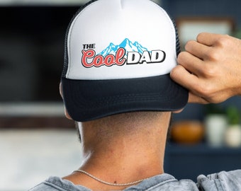 May include: A white and black trucker hat with a blue and white mountain graphic and the text "The Cool Dad" in red and blue lettering.