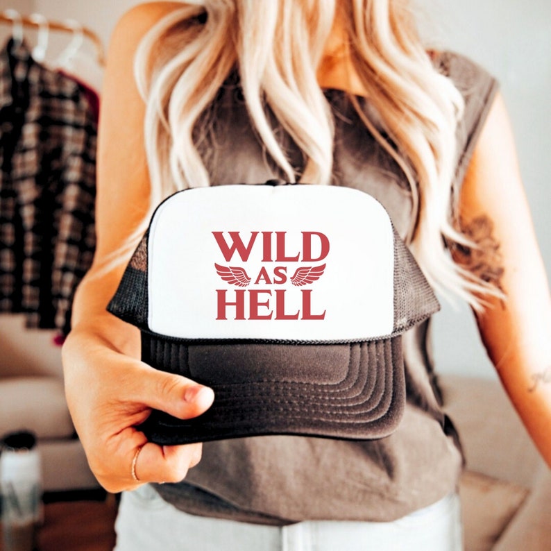 May include: A white and black trucker hat with the text "WILD AS HELL" in red lettering and red wings on either side of the text.