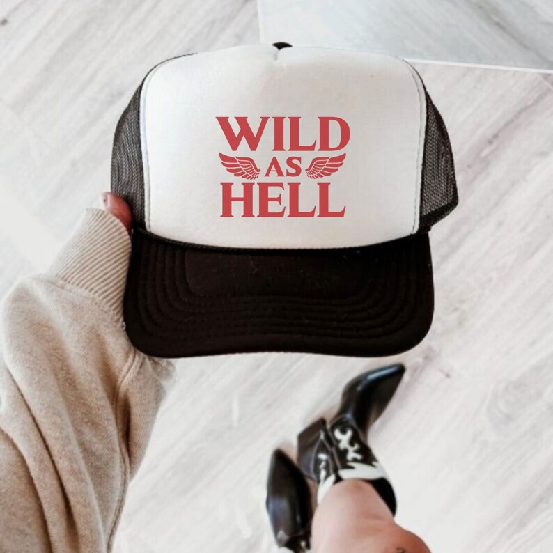 May include: A white trucker hat with a black mesh back. The front of the hat has red text that says "WILD AS HELL" with red wings on either side of the word "AS".