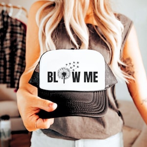 May include: A white and black trucker hat with a dandelion design and the text "BLOW ME" on the front.