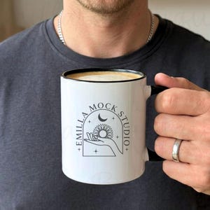 Op de afbeelding: Witte keramische mok met een zwarte handgreep en rand, gevuld met koffie. De mok heeft een zwart-witte afbeelding met de tekst "EMILLA MOCK STUDIO" en een ontwerp van een hand die een zonnebloem, een maan en sterren vasthoudt.