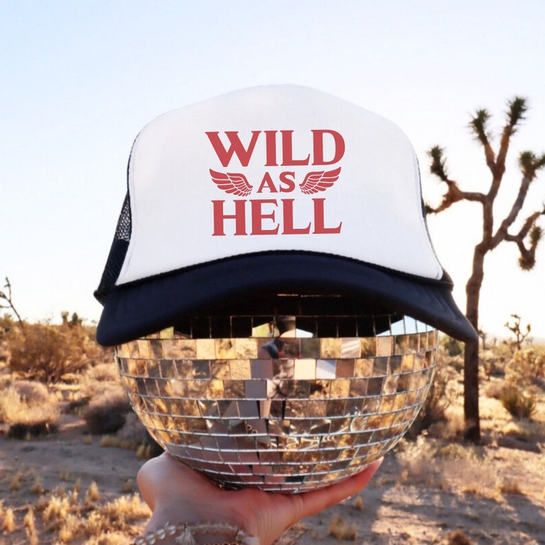 May include: White trucker hat with a black mesh back. The hat has red text that says "WILD AS HELL" with wings on either side of the word "AS".
