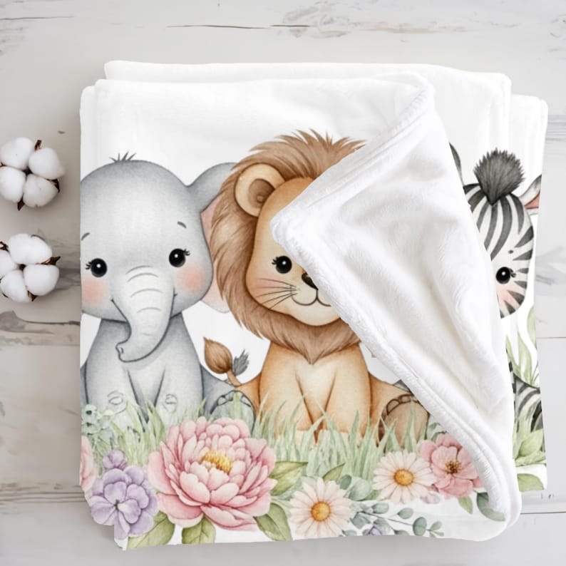 May include: White baby blanket featuring an elephant, lion, and zebra illustration. The blanket has a soft, plush texture and is decorated with colorful flowers and greenery. The blanket is folded, showing the design on the front.