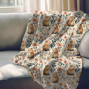 May include: A white throw blanket with a repeating pattern of brown guinea pigs and orange flowers. The guinea pigs are sitting up and have their paws in the air.