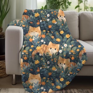 May include: A throw blanket featuring a repeating pattern of orange tabby cats nestled among green foliage and yellow and white flowers. The blanket is draped over a gray sofa, creating a cozy and inviting scene.