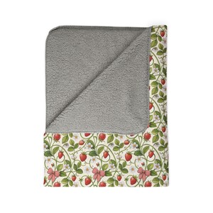 May include: A cozy blanket with a gray sherpa lining and a strawberry pattern. The design features red strawberries, green leaves, white flowers, and red gingham bows on a cream background. This blanket is perfect for snuggling.