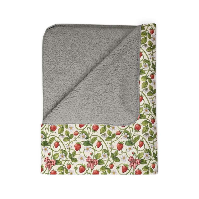 May include: A cozy blanket with a gray sherpa lining and a strawberry pattern. The design features red strawberries, green leaves, white flowers, and red gingham bows on a cream background. This blanket is perfect for snuggling.