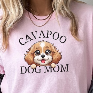 May include: A light pink t-shirt with the words "CAVAPOO DOG MOM" in black lettering. The shirt features a cartoon illustration of a Cavapoo dog with brown and white fur, big eyes, and a pink tongue.