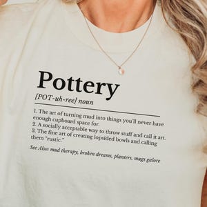 May include: A white t-shirt with a black graphic design that defines the word "Pottery" and includes a humorous definition of the craft. The definition reads: "The art of turning mud into things you'll never have enough cupboard space for. A socially acceptable way to throw stuff and call it art. The fine art of creating lopsided bowls and calling them "rustic." See Also: mud therapy, broken dreams, planters, mugs galore"