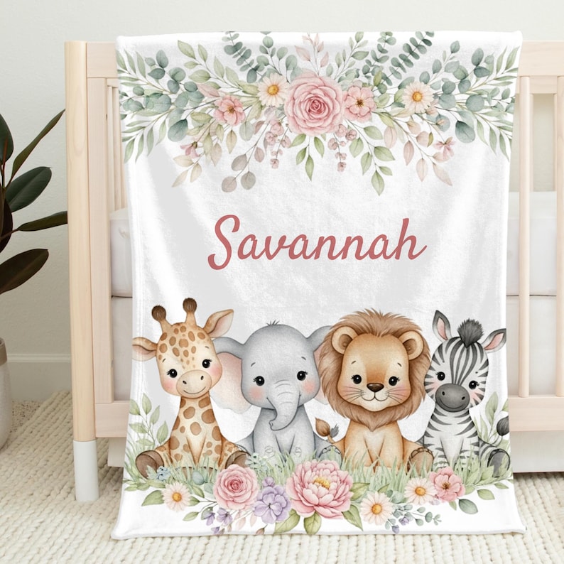 May include: White baby blanket featuring the name "Savannah" in pink script. The blanket showcases a watercolor design of a giraffe, elephant, lion, and zebra, surrounded by floral and foliage accents. The blanket is designed for a nursery.