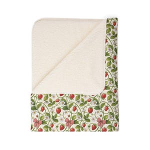 May include: A cream-colored blanket with a strawberry pattern. The blanket features a soft, plush interior and a decorative exterior with red strawberries, green leaves, white flowers, and red gingham bows. The blanket is folded over, revealing the plush interior.