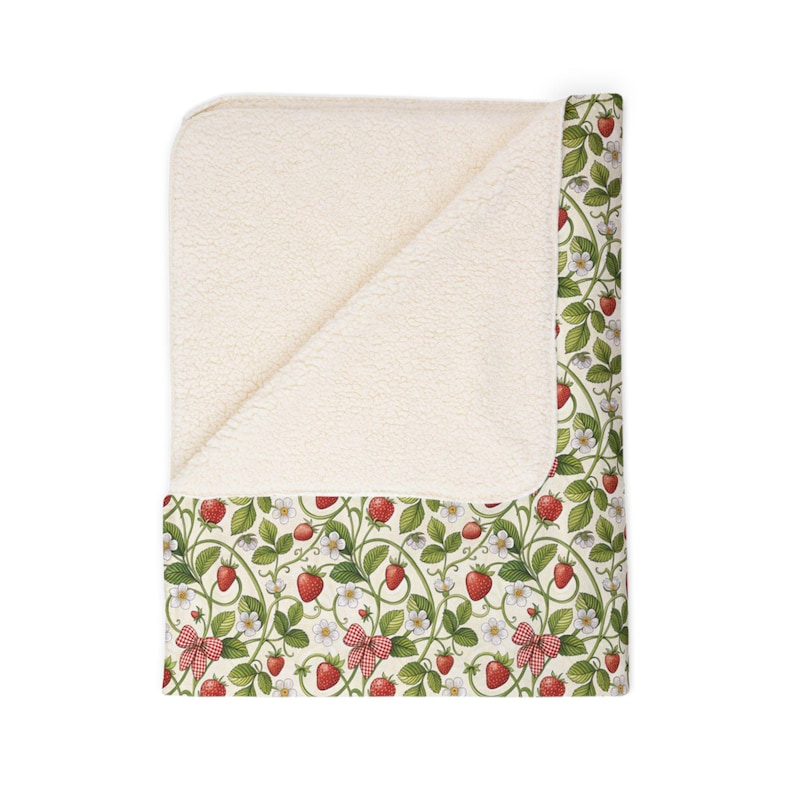 May include: A cream-colored blanket with a strawberry pattern. The blanket features a soft, plush interior and a decorative exterior with red strawberries, green leaves, white flowers, and red gingham bows. The blanket is folded over, revealing the plush interior.