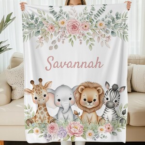 May include: White blanket featuring a floral design with the name "Savannah" in pink script. Below the text are illustrations of a giraffe, elephant, lion, and zebra. The blanket has a soft, inviting appearance.