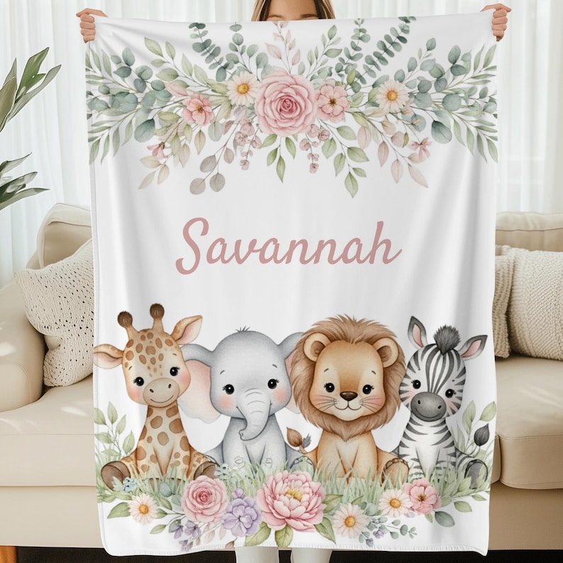 May include: White blanket featuring a floral design with the name "Savannah" in pink script. Below the text are illustrations of a giraffe, elephant, lion, and zebra. The blanket has a soft, inviting appearance.