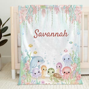 May include: A baby blanket featuring an underwater scene with a dolphin, octopus, turtle, seahorses, and jellyfish. The blanket is light blue with coral and seaweed accents. The name "Savannah" is written in red script across the top.