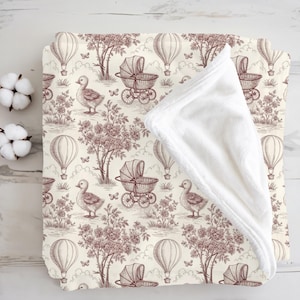May include: A cream-colored baby blanket with a repeating pattern of hot air balloons, ducklings, and vintage baby carriages in a sepia tone. The blanket has a soft, white, plush backing.