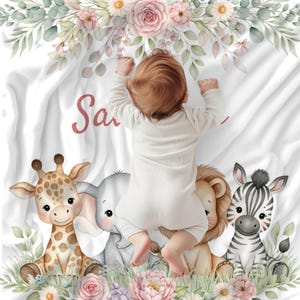 May include: A white baby blanket with the name "Sa" in pink script, featuring a baby lying on top. The blanket is decorated with a giraffe, elephant, lion, and zebra, surrounded by flowers and greenery.