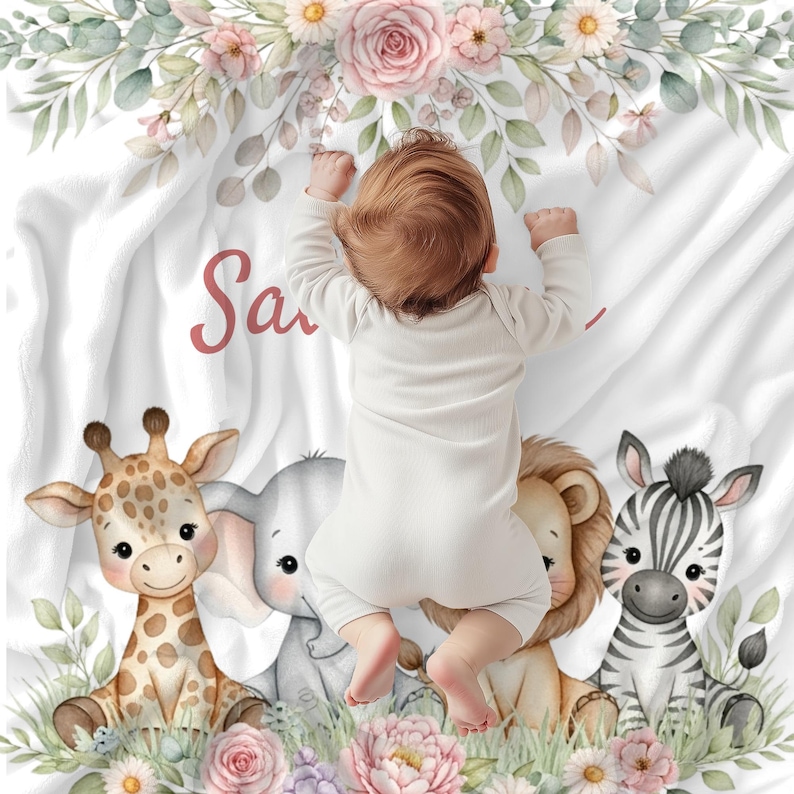 May include: A white baby blanket with the name "Sa" in pink script, featuring a baby lying on top. The blanket is decorated with a giraffe, elephant, lion, and zebra, surrounded by flowers and greenery.