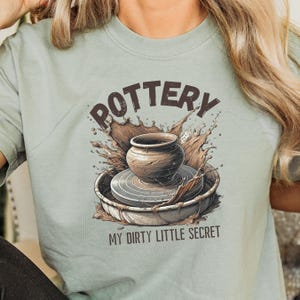 May include: A light blue t-shirt with a brown pottery wheel and a clay pot with the text "POTTERY MY DIRTY LITTLE SECRET" printed on it.