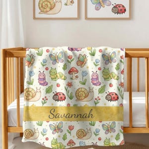 May include: A cream-colored baby blanket with a repeating pattern of colorful butterflies, snails, ladybugs, caterpillars, and mushrooms. The blanket has a gold band with the name "Savannah" printed on it. The blanket is draped over a wooden crib.