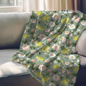 May include: A green throw blanket with a repeating pattern of yellow frogs, pink and white flowers, and green leaves.