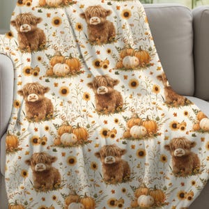 May include: A cream-coloured blanket with a repeating pattern of brown Highland cows, pumpkins, and sunflowers. The pumpkins are orange and white, and the sunflowers are yellow. The blanket is draped over a grey sofa.