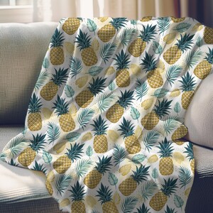 May include: A white throw blanket with a repeating pattern of golden pineapples and green and yellow leaves. The blanket is draped over a gray sofa, creating a cozy and tropical aesthetic. The blanket is made of a soft material.