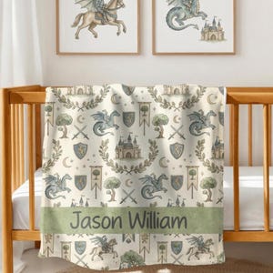 May include: A cream-colored baby blanket with a medieval theme, featuring dragons, castles, shields, and trees. The blanket has a green band with the name "Jason William" printed on it. The blanket is draped over a wooden crib.