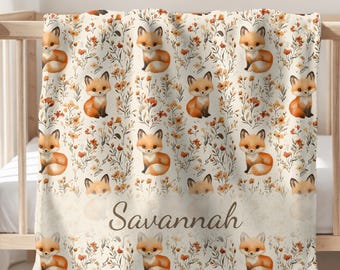 May include: A cream-colored baby blanket featuring a repeating pattern of watercolor foxes and floral elements in shades of orange, brown, and green. The name "Savannah" is printed in a cursive font across the bottom.