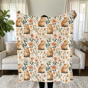 May include: A white fleece blanket with a repeating pattern of brown capybaras and orange flowers. The capybaras are standing on their hind legs and have their front paws raised.
