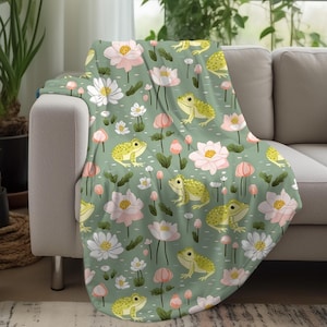 May include: A green throw blanket with a repeating pattern of pink flowers, green leaves, and yellow frogs. The blanket is soft and cozy, perfect for adding a touch of whimsy to any room.