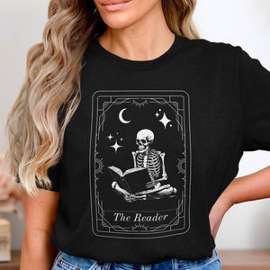 以下が含まれることがあります： 黒いTシャツに、本を読んでいる骸骨の白いグラフィックがプリントされています。グラフィックはタロットカード風のフレームの中にあり、「The Reader」というテキストが書かれています。