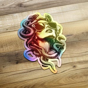 以下が含まれることがあります： サングラスをかけたメデューサとヘビのスタイリッシュなイラストが描かれたホログラフィックステッカー。虹色のイリデセント効果があり、木製の表面に配置されています。