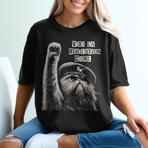 May include: A black short-sleeved t-shirt featuring a white graphic of a cat wearing a beret with a small dinosaur icon on it. The cat has one paw raised in the air. The text above the cat reads VIVA LA REVOLUTION CARL in a blocky font.
