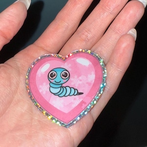 May include: A heart-shaped sticker with a pink background and a cartoon worm illustration. The worm is blue with a black outline and a smiling face. The sticker has a glittery, iridescent border and snowflake accents.