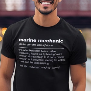 May include: A black t-shirt with white text that reads "marine mechanic" and a definition of the term. The person wearing the shirt is standing on a wooden dock.